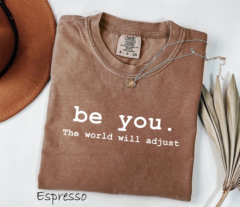 K&ouml;nnte beinhalten: Espressofarbenes T-Shirt mit dem wei&szlig;en Text "be you. The world will adjust". Das Shirt hat einen Rundhalsausschnitt und ein kleines Etikett am Ausschnitt. Eine silberne Halskette mit Anh&auml;nger liegt &uuml;ber dem Shirt.
