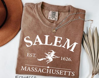 May include: A brown short-sleeved t-shirt featuring white screen-printed text that reads SALEM EST. 1626 MASSACHUSETTS with a graphic of a witch flying on a broomstick. The shirt has a crew neckline and a Comfort Colors brand label inside the collar.