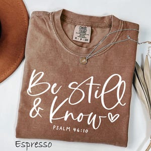 May include: An espresso-colored short-sleeve t-shirt with a crew neckline. The shirt features white screen-printed text that reads "Be still & Know" followed by "PSALM 46:10" and a small heart icon. The interior neck label reads "Comfort Colors".