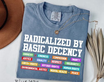 May include: A blue jean colored t-shirt with white text that reads RADICALIZED BY BASIC DECENCY. Below the text are colorful rectangular blocks with words including EMPATHY, KINDNESS, COMPASSION, DIGNITY, JUSTICE, EQUALITY, RESPECT, SOLIDARITY, LGBTQ+ RIGHTS, REFUGEE RIGHTS, HUMAN RIGHTS, ENVIRONMENTAL JUSTICE, MENTAL HEALTH, and PEACE.