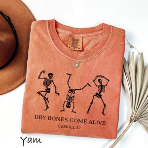 Peut inclure: Un t-shirt à manches courtes de couleur terre cuite avec un motif noir représentant trois squelettes en train de danser. Sous les squelettes, le texte indique "DRY BONES COME ALIVE" et "EZEKIEL 37". Le t-shirt a un col rond et une étiquette Comfort Colors.