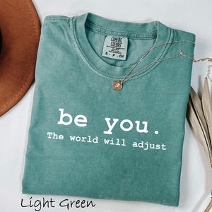 K&ouml;nnte beinhalten: Hellgr&uuml;nes T-Shirt mit dem wei&szlig;en Text "be you. The world will adjust". Eine goldene Halskette mit Anh&auml;nger liegt auf dem Shirt. Das Shirt ist gefaltet und hat ein Comfort Colors-Etikett.