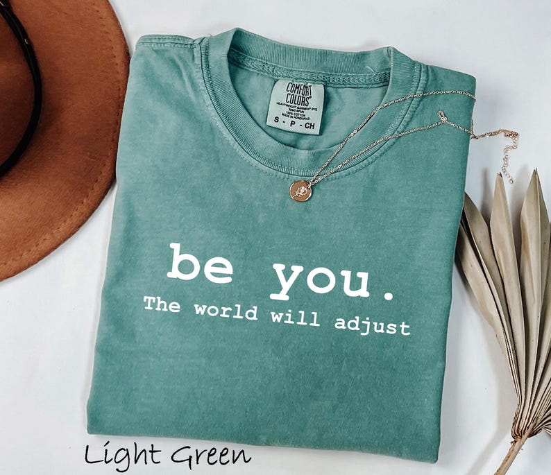 K&ouml;nnte beinhalten: Hellgr&uuml;nes T-Shirt mit dem wei&szlig;en Text "be you. The world will adjust". Eine goldene Halskette mit Anh&auml;nger liegt auf dem Shirt. Das Shirt ist gefaltet und hat ein Comfort Colors-Etikett.