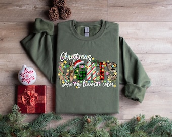 May include: An olive green crewneck sweatshirt featuring a graphic print that reads Christmas LIGHTS Are my favorite color. The letters are filled with various patterns including plaid, leopard print, and candy cane stripes, accented by string lights.
