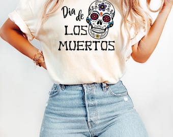 May include: A cream-colored short-sleeve t-shirt featuring a graphic of a sugar skull decorated with purple, red, and blue floral patterns. The text "Dia de LOS MUERTOS" is printed in black, with the word "LOS" stylized to resemble bone segments.