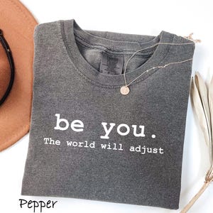 K&ouml;nnte beinhalten: Ein graues T-Shirt mit den Worten "be you. The world will adjust" in Wei&szlig; gedruckt. Eine goldene Halskette mit einem kreisf&ouml;rmigen Anh&auml;nger liegt auf dem Shirt. Ein brauner Hut ist in der oberen linken Ecke zu sehen.