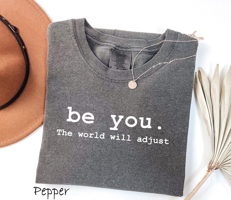 K&ouml;nnte beinhalten: Ein graues T-Shirt mit den Worten "be you. The world will adjust" in Wei&szlig; gedruckt. Eine goldene Halskette mit einem kreisf&ouml;rmigen Anh&auml;nger liegt auf dem Shirt. Ein brauner Hut ist in der oberen linken Ecke zu sehen.