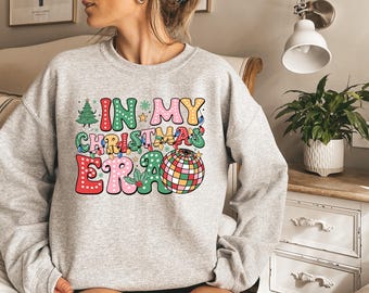 May include: A light gray crewneck sweatshirt featuring a colorful graphic print that reads "IN MY CHRISTMAS ERA" in bold, bubbly letters. The design includes a decorated Christmas tree, a disco ball, holly leaves, and small string lights.