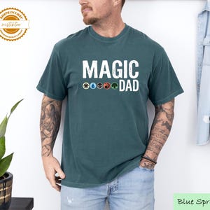 May include: A blue spruce t-shirt with the text "MAGIC DAD" in white block letters. Below the text are five colored symbols. The shirt is a crew neck and is worn by a person.