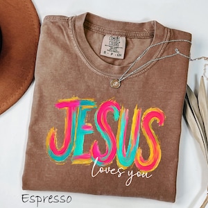 Puede incluir: Camiseta color espresso con las palabras "JESUS loves you" en vibrantes y coloridas pinceladas. La camiseta tiene cuello redondo y una peque&ntilde;a etiqueta en el cuello. Un collar plateado con un colgante est&aacute; drapeado sobre la camiseta.