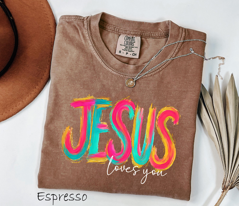 Puede incluir: Camiseta color espresso con las palabras "JESUS loves you" en vibrantes y coloridas pinceladas. La camiseta tiene cuello redondo y una peque&ntilde;a etiqueta en el cuello. Un collar plateado con un colgante est&aacute; drapeado sobre la camiseta.