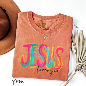 Puede incluir: Una camiseta de color coral con las palabras "JESUS loves you" en un dise&ntilde;o colorido de pinceladas. La camiseta tiene cuello redondo y mangas cortas. Un collar de oro con un colgante est&aacute; drapeado sobre la camiseta. Un sombrero marr&oacute;n est&aacute; en el fondo.