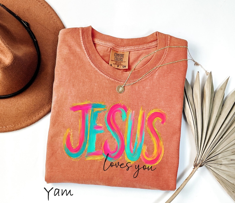 Puede incluir: Una camiseta de color coral con las palabras "JESUS loves you" en un dise&ntilde;o colorido de pinceladas. La camiseta tiene cuello redondo y mangas cortas. Un collar de oro con un colgante est&aacute; drapeado sobre la camiseta. Un sombrero marr&oacute;n est&aacute; en el fondo.