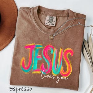 May include: A brown short-sleeved t-shirt with a crew neckline and a Comfort Colors brand label. The front features the word JESUS in large, multi-colored brushstroke lettering in pink, yellow, and teal, with the white script text loves you below.