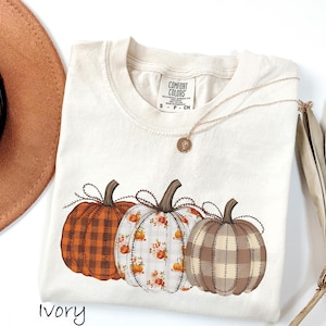 May include: An ivory-coloured t-shirt with three decorative pumpkins. The pumpkins have stitched details and different patterns: plaid, floral, and brown and tan plaid. A gold necklace and a brown hat are also visible.