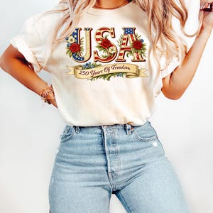 Może przedstawiać: Kremowa koszulka z napisem "USA" w patriotycznym wzornictwie. Litery zdobione są czerwonymi r&oacute;żami, niebieskimi kwiatami i wzorami amerykańskiej flagi. Poniżej widnieje napis "250 Years Of Freedom".