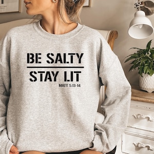May include: A light gray crewneck sweatshirt featuring black text that reads BE SALTY STAY LIT with the reference MATT 5:13-14 printed below. The garment has long sleeves, a ribbed neckline, and a standard fit, showcasing a clear graphic design.