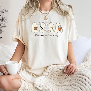 May include: An ivory short-sleeve t-shirt featuring a graphic of four cartoon ghosts engaged in various activities, including reading and drinking coffee. Below the ghosts, the text reads "Para normal activities" in a brown, cursive font.
