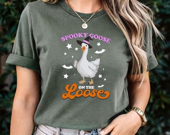 May include: A dark olive green short-sleeve t-shirt featuring a graphic of a white goose wearing a black witch hat. The text reads "SPOOKY GOOSE" in purple and "ON THE LOOSE" in orange, surrounded by white stars and small black bat silhouettes.