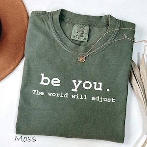 K&ouml;nnte beinhalten: Olivgr&uuml;nes T-Shirt mit dem wei&szlig;en Text "be you. The world will adjust". Eine goldene Halskette mit Anh&auml;nger liegt auf dem Shirt. Ein brauner Hut und getrocknete Palmbl&auml;tter sind im Hintergrund.