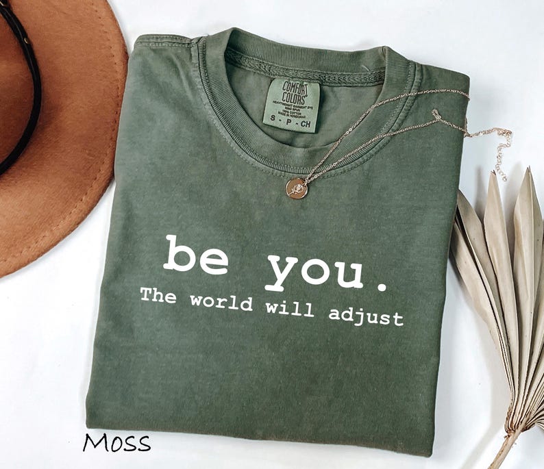 K&ouml;nnte beinhalten: Olivgr&uuml;nes T-Shirt mit dem wei&szlig;en Text "be you. The world will adjust". Eine goldene Halskette mit Anh&auml;nger liegt auf dem Shirt. Ein brauner Hut und getrocknete Palmbl&auml;tter sind im Hintergrund.