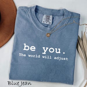 K&ouml;nnte beinhalten: Blaues Jeans-T-Shirt mit dem wei&szlig;en Text "be you. The world will adjust". Eine goldene Halskette mit Anh&auml;nger liegt &uuml;ber dem Shirt. Ein brauner Hut und eine getrocknete Pflanze befinden sich im Hintergrund.