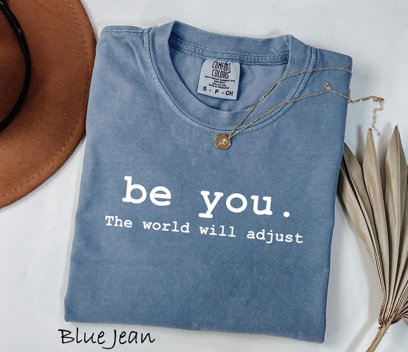 K&ouml;nnte beinhalten: Blaues Jeans-T-Shirt mit dem wei&szlig;en Text "be you. The world will adjust". Eine goldene Halskette mit Anh&auml;nger liegt &uuml;ber dem Shirt. Ein brauner Hut und eine getrocknete Pflanze befinden sich im Hintergrund.