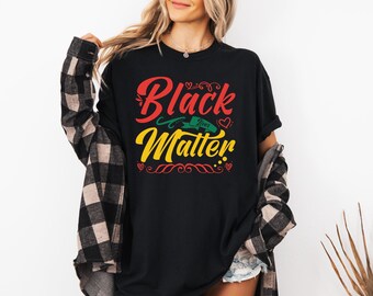 May include: A black short-sleeved t-shirt featuring the red text Black and yellow text Matter with the words Lives in a green banner. The design includes small red hearts and decorative flourishes. A black and beige plaid flannel shirt is worn over it.