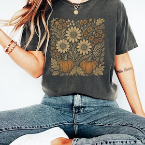 May include: A charcoal gray short-sleeve t-shirt featuring a square graphic print of two orange pumpkins surrounded by gold-toned line art illustrations of daisies, autumn leaves, and a pinecone, paired with blue denim jeans and white sneakers.