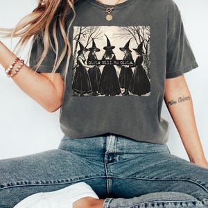 May include: A charcoal gray short-sleeve t-shirt featuring a graphic print of five figures in pointed hats and long dresses standing in a circle. The text "Girls Will Be Girls." is printed in white across the center of the graphic illustration.