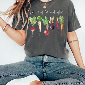 May include: A charcoal gray short-sleeve t-shirt featuring a watercolor-style graphic of various root vegetables including radishes, beets, and carrots. Above the vegetables, the white text reads, Let's root for each other.