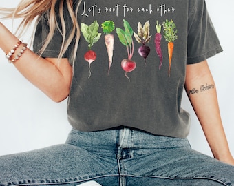 May include: A charcoal gray short-sleeve t-shirt featuring a watercolor-style graphic of various root vegetables including radishes, beets, and carrots. Above the vegetables, the white text reads, Let's root for each other.