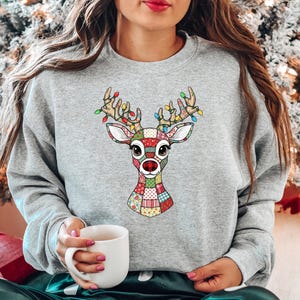 May include: A heather gray crewneck sweatshirt featuring a central graphic of a reindeer head. The reindeer is designed with a colorful patchwork pattern and has antlers decorated with multi-colored Christmas light bulbs.