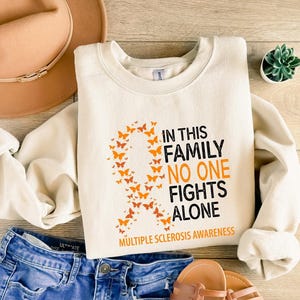 May include: A cream-colored crewneck sweatshirt featuring an orange awareness ribbon composed of small butterflies. The text reads: IN THIS FAMILY NO ONE FIGHTS ALONE, with MULTIPLE SCLEROSIS AWARENESS printed in orange at the bottom.