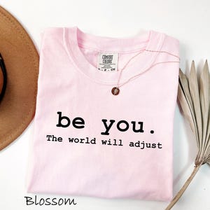 K&ouml;nnte beinhalten: Ein hellrosa T-Shirt mit den Worten "be you. The world will adjust" in Schwarz gedruckt. Eine goldene Halskette mit einem kleinen Anh&auml;nger liegt auf dem Shirt. Ein brauner Hut ist in der oberen linken Ecke zu sehen.