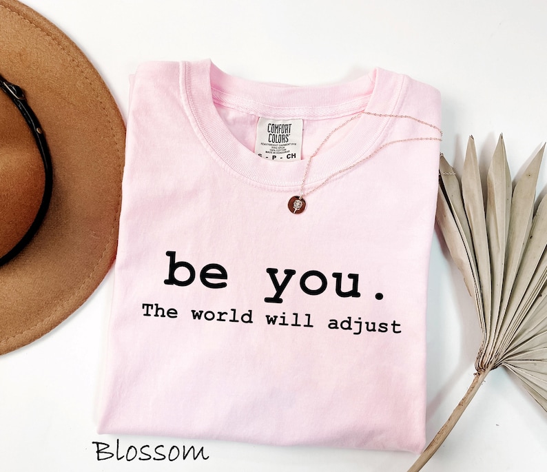 K&ouml;nnte beinhalten: Ein hellrosa T-Shirt mit den Worten "be you. The world will adjust" in Schwarz gedruckt. Eine goldene Halskette mit einem kleinen Anh&auml;nger liegt auf dem Shirt. Ein brauner Hut ist in der oberen linken Ecke zu sehen.