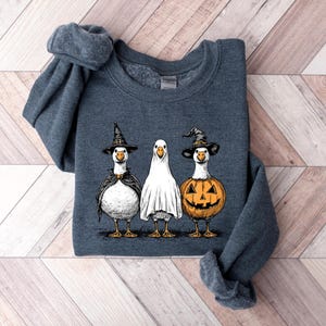 May include: A dark heather gray crewneck sweatshirt featuring a graphic print of three white geese dressed for Halloween. One goose wears a black witch hat and cape, the middle goose is draped in a white ghost sheet, and the third wears a pumpkin costume.