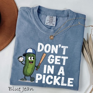 May include: Blue jean colored t-shirt with the phrase "DON'T GET IN A PICKLE" in white block letters. A cartoon pickle wearing a baseball cap and holding a bat and baseball is featured on the front. A gold necklace is draped across the shirt.