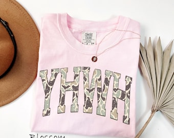 May include: A light pink t-shirt featuring the text YHWH in a block font with a camouflage pattern of green, brown, and cream. The shirt has a Comfort Colors label at the neck and is displayed alongside a brown hat and a dried palm leaf.