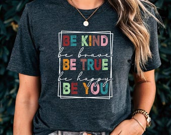 May include: A dark charcoal gray short-sleeve t-shirt featuring a white rectangular frame containing the text BE KIND, be brave, BE TRUE, be happy, BE YOU in a mix of colorful, distressed block and script fonts with red, blue, and yellow.