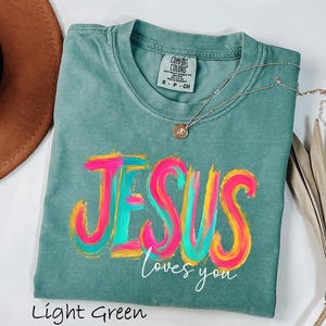 Puede incluir: Camiseta verde claro con la frase "JESUS loves you" en un dise&ntilde;o colorido de pincelada. La camiseta tiene cuello redondo y mangas cortas. Un collar plateado con un peque&ntilde;o colgante est&aacute; sobre la camiseta.