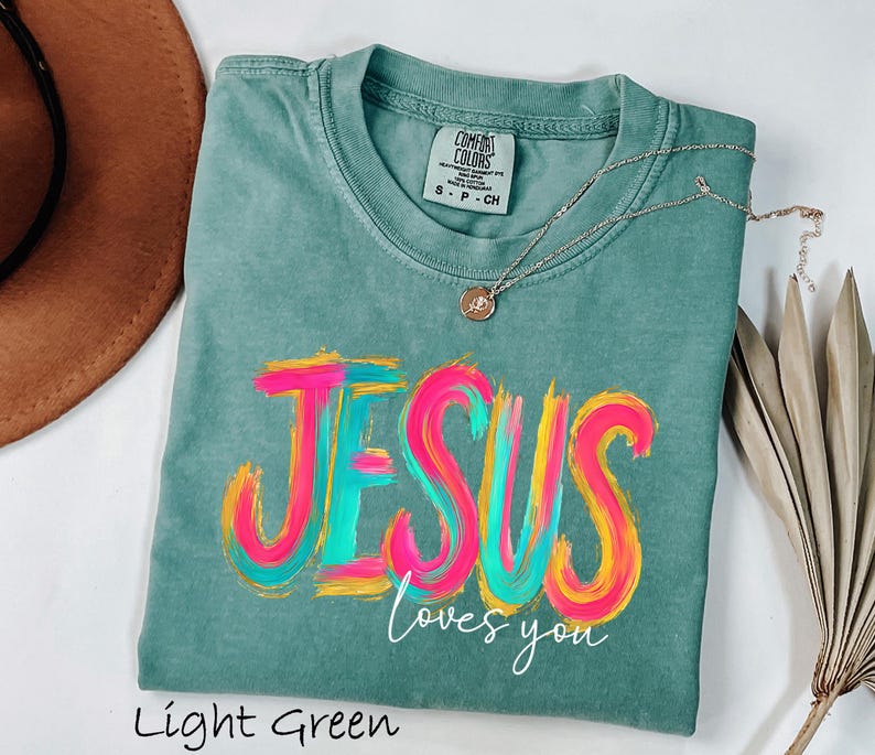 Puede incluir: Camiseta verde claro con la frase "JESUS loves you" en un dise&ntilde;o colorido de pincelada. La camiseta tiene cuello redondo y mangas cortas. Un collar plateado con un peque&ntilde;o colgante est&aacute; sobre la camiseta.