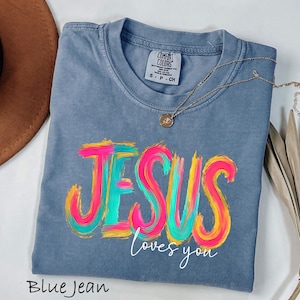 Puede incluir: Camiseta azul vaquero con las palabras "JESUS loves you" en un dise&ntilde;o colorido de pinceladas. Un collar dorado con un colgante descansa sobre la camiseta. La camiseta est&aacute; etiquetada como "Comfort Colors".