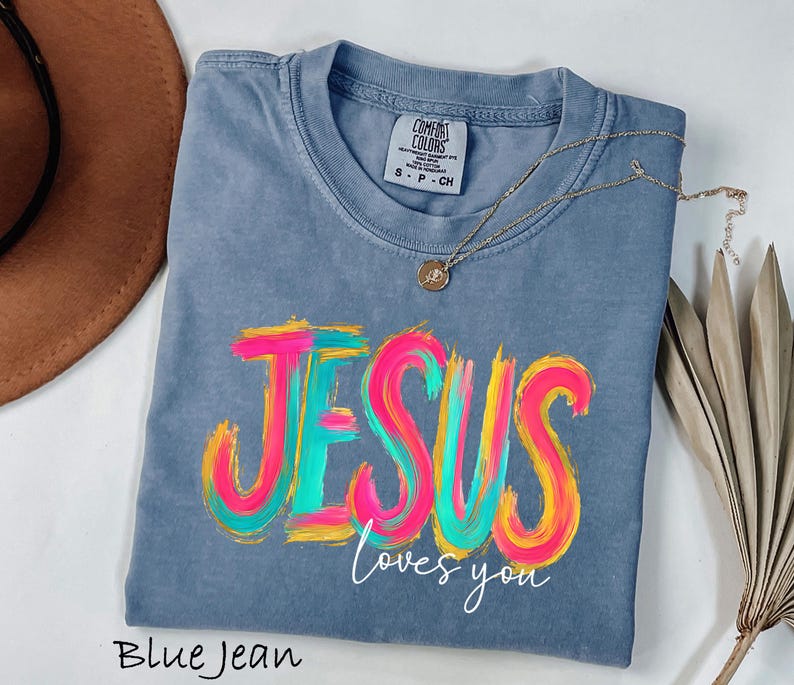 Puede incluir: Camiseta azul vaquero con las palabras "JESUS loves you" en un dise&ntilde;o colorido de pinceladas. Un collar dorado con un colgante descansa sobre la camiseta. La camiseta est&aacute; etiquetada como "Comfort Colors".