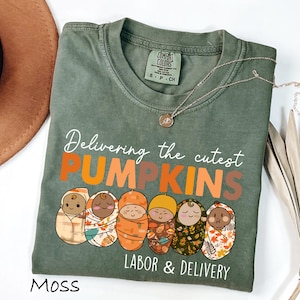 May include: Moss green t-shirt with the text "Delivering the cutest PUMPKINS LABOR & DELIVERY". The shirt features a graphic of five swaddled babies in various patterns and colors. A gold necklace is draped on the shirt.