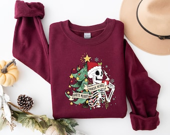 May include: A maroon crewneck sweatshirt featuring a graphic print of a split design. One side shows a decorated Christmas tree with yellow stars, while the other side depicts a skeleton wearing a Santa hat and holding a mug, with the text "SORTA MERRY" and "SORTA SCARY" on banners.