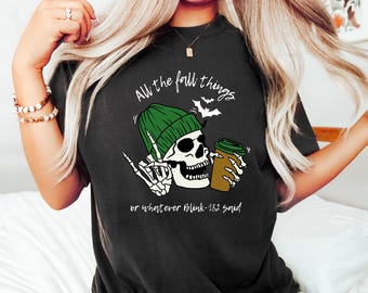 May include: A black t-shirt featuring a graphic of a skeleton wearing a green beanie, holding a brown coffee cup, and making a hand gesture. The text reads "All the fall things" and "or whatever Blink-182 said" with small white bat illustrations.