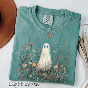 May include: A light green short-sleeve t-shirt with a crew neckline and a Comfort Colors label. The graphic features a white ghost standing in a field of colorful wildflowers. A thin silver chain necklace with a small round pendant rests on the shirt.