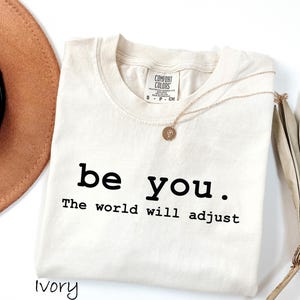 K&ouml;nnte beinhalten: Elfenbeinfarbenes T-Shirt mit dem schwarzen Text "be you. The world will adjust". Eine goldene Halskette mit Anh&auml;nger liegt auf dem Shirt. Ein brauner Hut und getrocknete Palmbl&auml;tter sind im Hintergrund.