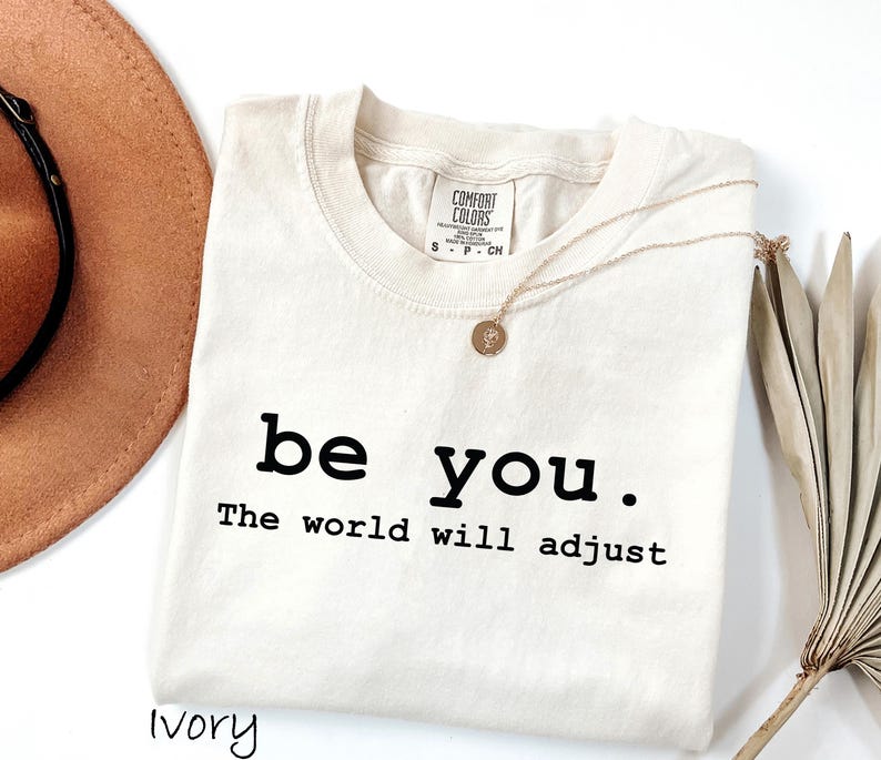 K&ouml;nnte beinhalten: Elfenbeinfarbenes T-Shirt mit dem schwarzen Text "be you. The world will adjust". Eine goldene Halskette mit Anh&auml;nger liegt auf dem Shirt. Ein brauner Hut und getrocknete Palmbl&auml;tter sind im Hintergrund.